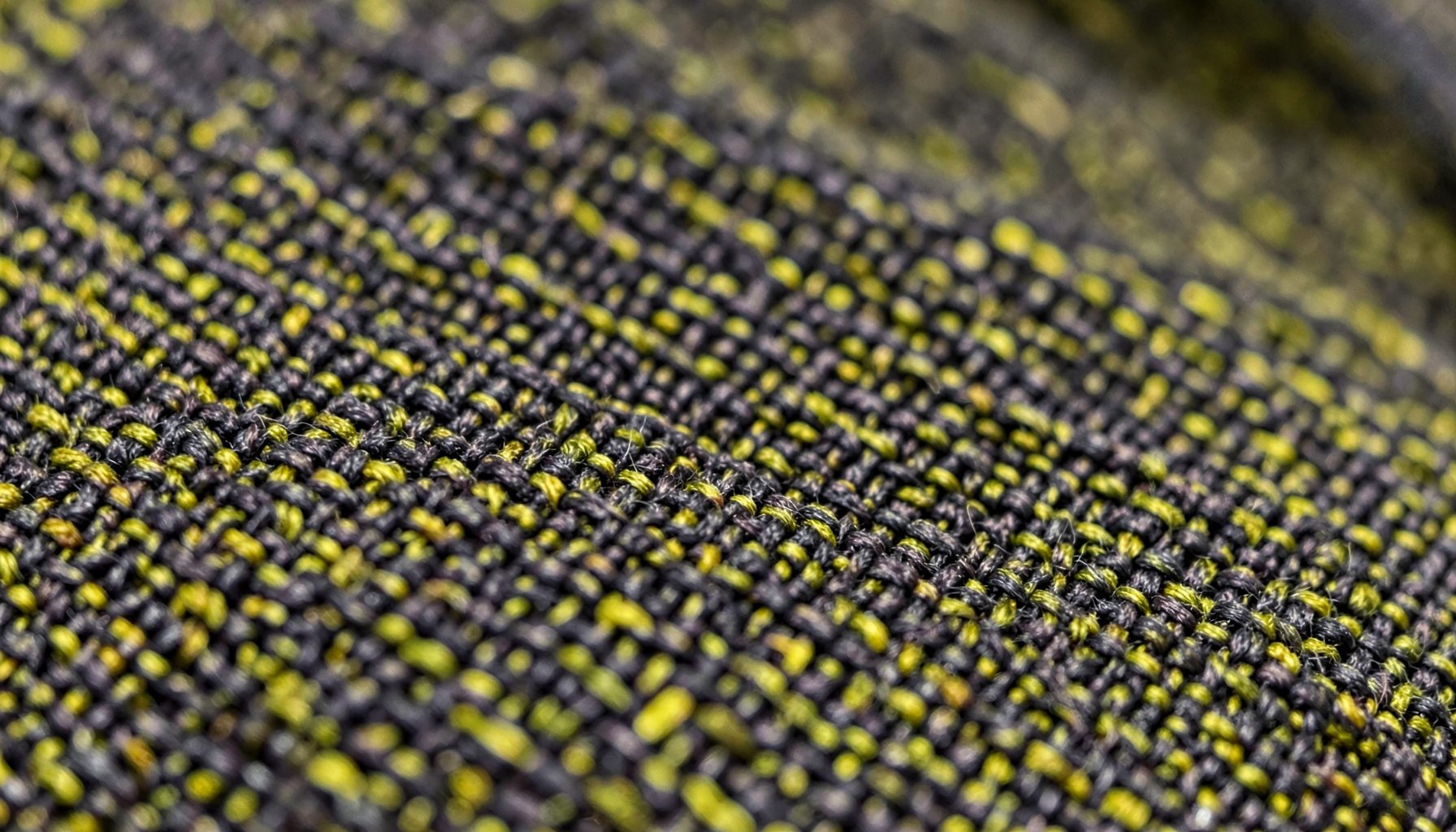 Extreme close-up of a textured woven fabric in warm charcoal and muted olive tones, with subtle highlights catching the thread ridges.