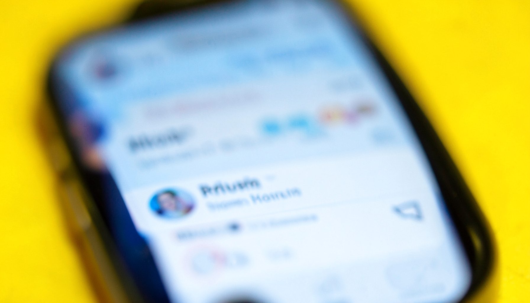 Extreme close-up photograph of a smartphone screen showing a social media feed with soft blue and white interface tones, blurred in the background, conveying a sense of scrolling through updates.