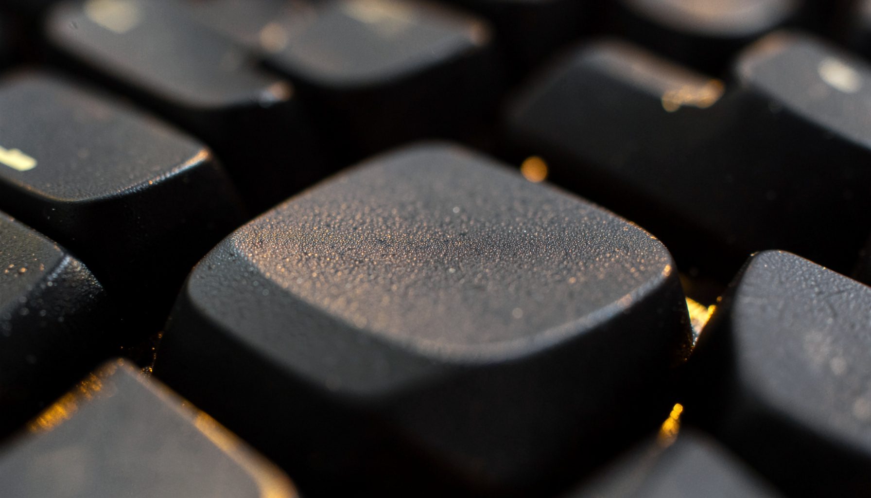 Extreme close-up photograph of matte black keyboard keycaps with subtle backlight glow, deep charcoal tones and soft amber highlights